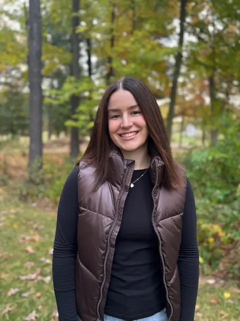 Alyssa with a brown fall vest standing outside with trees and fall leaves in the background